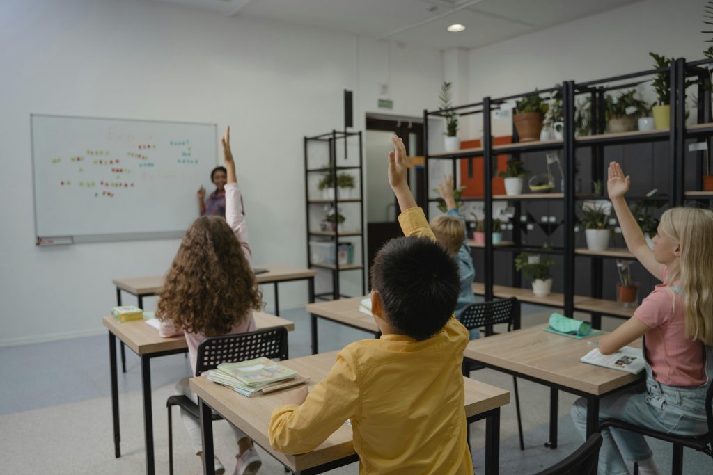A classroom of students in specialist education, with their hands up to answer a question. The teacher is at the front of the class talking to students.